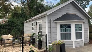 Une petite maison de plain-pied, à l’extérieur gris et au toit noir. Elle possède un jardin clôturé à l’arrière, avec un espace patio, plusieurs plantes en pot et un petit potager.