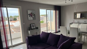 A modern living room with a purple sofa, a glass coffee table, and a dining area with a white table and chairs. - Apartment ST JEAN Centre Ville Wifi Balcony Parking (Saint-Jean-de-Monts)