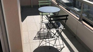 A balcony with a table and chairs, a green bench, and a view of buildings and a car in the background. - Apartment ST JEAN Centre Ville Wifi Balcony Parking (Saint-Jean-de-Monts)