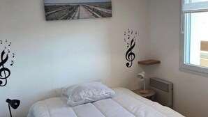 A neatly made bed with white bedding, a small bedside table with a lamp, a wall-mounted picture, and a window with blinds. - Apartment ST JEAN Centre Ville Wifi Balcony Parking (Saint-Jean-de-Monts)
