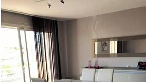 A modern dining area with a glass table and white chairs, a kitchen with white cabinets, and a large window with curtains. - Apartment ST JEAN Centre Ville Wifi Balcony Parking (Saint-Jean-de-Monts)