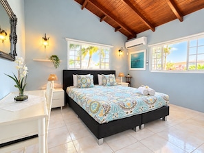 A bedroom with a bed, bedside tables, a mirror, and a window with a view of palm trees.