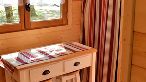 Puisesta materiaalista rakennettu mökkimäinen huone, jossa on puinen pöytä, kaksi laatikkoa ja ikkuna, jossa on pitsiverhot.