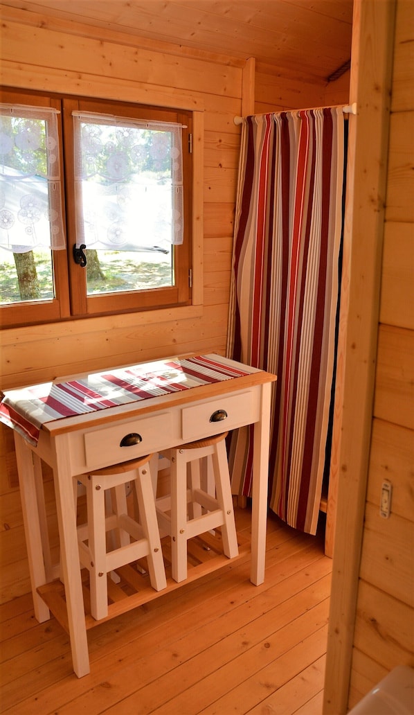 A wooden cabin room with a wooden table, two drawers, and a window with lace curtains.