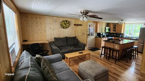 Un salon confortable avec un canapé, un pouf et une table basse, une cuisine dotée de meubles en bois et d’un four à micro-ondes, et un ventilateur de plafond.