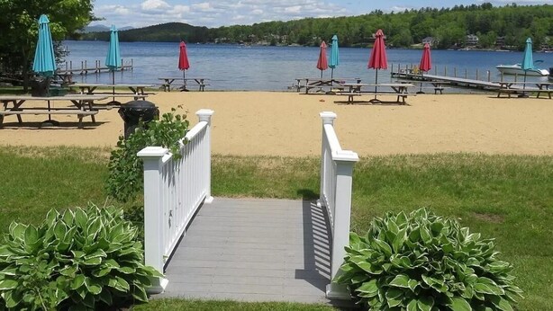 Une plage avec des tables de pique-nique et des parasols, une terrasse en bois et une vue sur un lac.