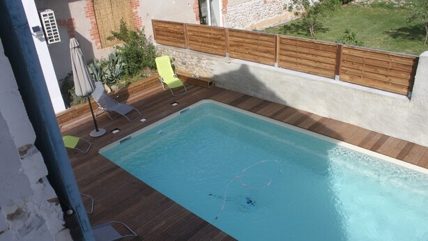 A rectangular swimming pool with a wooden deck, surrounded by a low stone wall and a wooden fence. There are two lounge chairs and a small umbrella. - Le Clos de L'arc (Vallon Pont d'Arc)