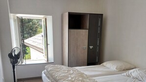 A bedroom with a bed, a wardrobe, a standing fan, and a window with a view of a building. - Le Clos de L'arc (Vallon Pont d'Arc)