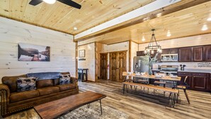 A spacious living room with a wooden ceiling, a large sofa, a coffee table, and a dining area with a bench and table. - Cabin with hot tub close to all the action! (Pigeon Forge)
