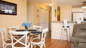 Een eetgedeelte met een ronde glazen tafel en witte stoelen, een keuken met witte kasten, en een plafondventilator.
