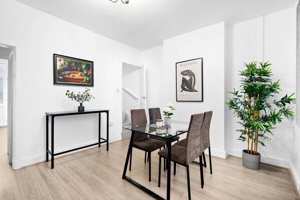 A modern dining area with a glass table, black metal frame, and brown upholstered chairs. There is a potted plant and a framed artwork on the wall.