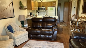 Een woonkamer met een leren bank, een fauteuil met patroon en een salontafel. Achtergrond is een keuken met houten kasten en apparatuur.