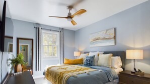 A bedroom with a bed, bedside lamps, a ceiling fan, a window with curtains, a painting on the wall, and a desk with a plant.