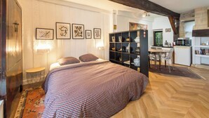 A bedroom with a bed, a small table, a bookshelf, and a kitchen area in the background.