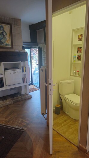 A room with a white radiator, a bookshelf, a door leading to a bathroom with a toilet and a shelf with toiletries.