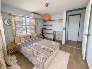 A bedroom with a patterned bedspread, a bedside table with a lamp, a hanging light fixture, a wardrobe, and a door with a blue frame.