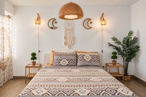 A bedroom with a patterned bedspread, two bedside tables with plants, a large woven pendant light, wall decorations including a crescent moon and a dreamcatcher, and a large potted plant.