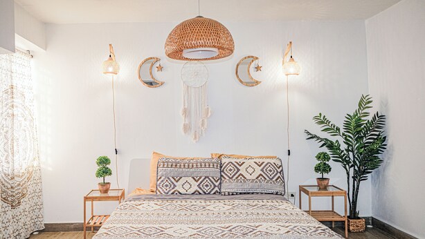 A bedroom with a patterned bedspread, two bedside tables with plants, a hanging woven light fixture, and wall decorations including a dreamcatcher and crescent moons.