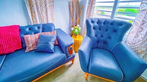 A room with a blue sofa and armchair, patterned curtains, and a view of greenery outside.