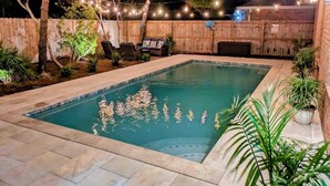A rectangular swimming pool with a tiled deck, surrounded by a wooden fence and string lights. - Heated Pool • Steps to Beach w/ Private Access (Miramar Beach)
