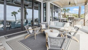 Une terrasse extérieure couverte, aménagée avec des sièges, un barbecue et de grandes fenêtres offrant une vue sur l’océan.