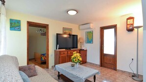 A living room with a sofa, a coffee table with a vase of flowers, a television on a wooden cabinet, and a door leading to another room.