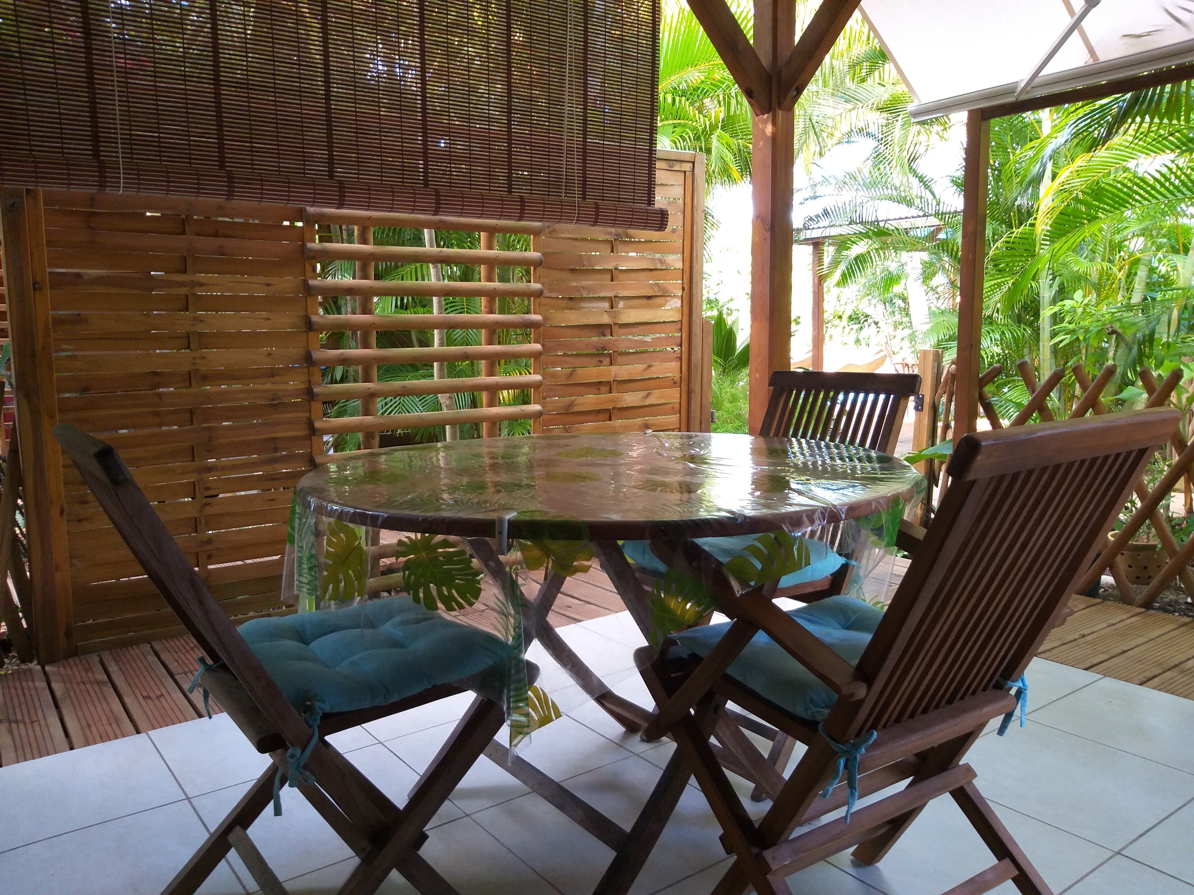 A covered outdoor seating area with a round table and wooden chairs, surrounded by a bamboo fence and lush greenery.