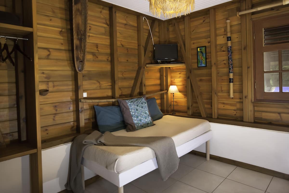 A wooden cabin room with a bed, a television on a shelf, a lamp, and a window with blinds.
