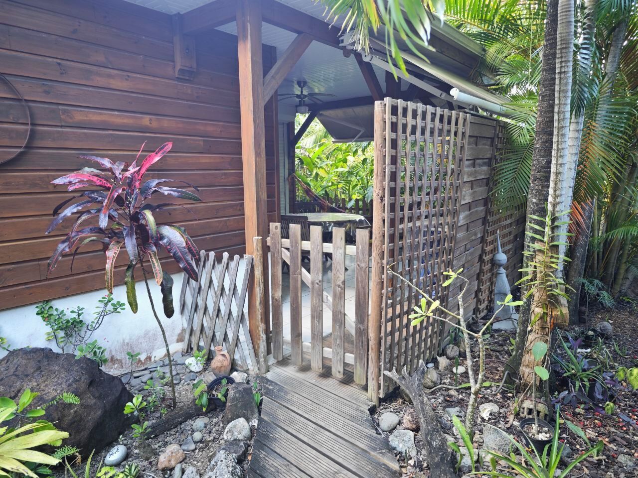 A wooden gate leading to a covered porch with a ceiling fan, surrounded by tropical plants and a small garden area.