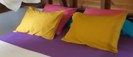Uma cama com colchão roxo e duas almofadas, uma amarela e outra rosa, em um quarto com paredes de madeira.