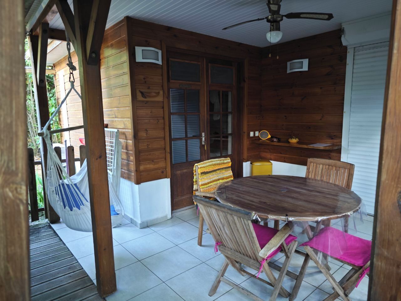 A wooden patio with a round table and chairs, a hammock, and a ceiling fan.
