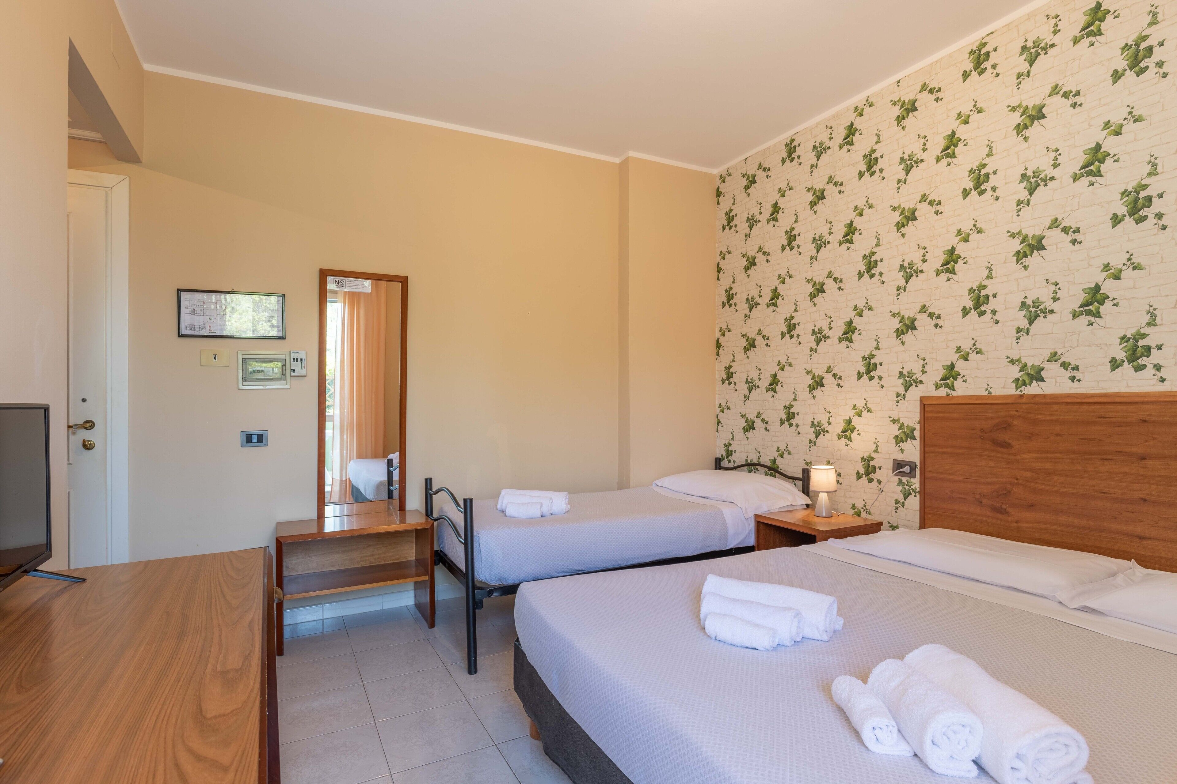 A hotel room with two single beds, a wooden desk, a mirror, and a wall with a floral pattern.