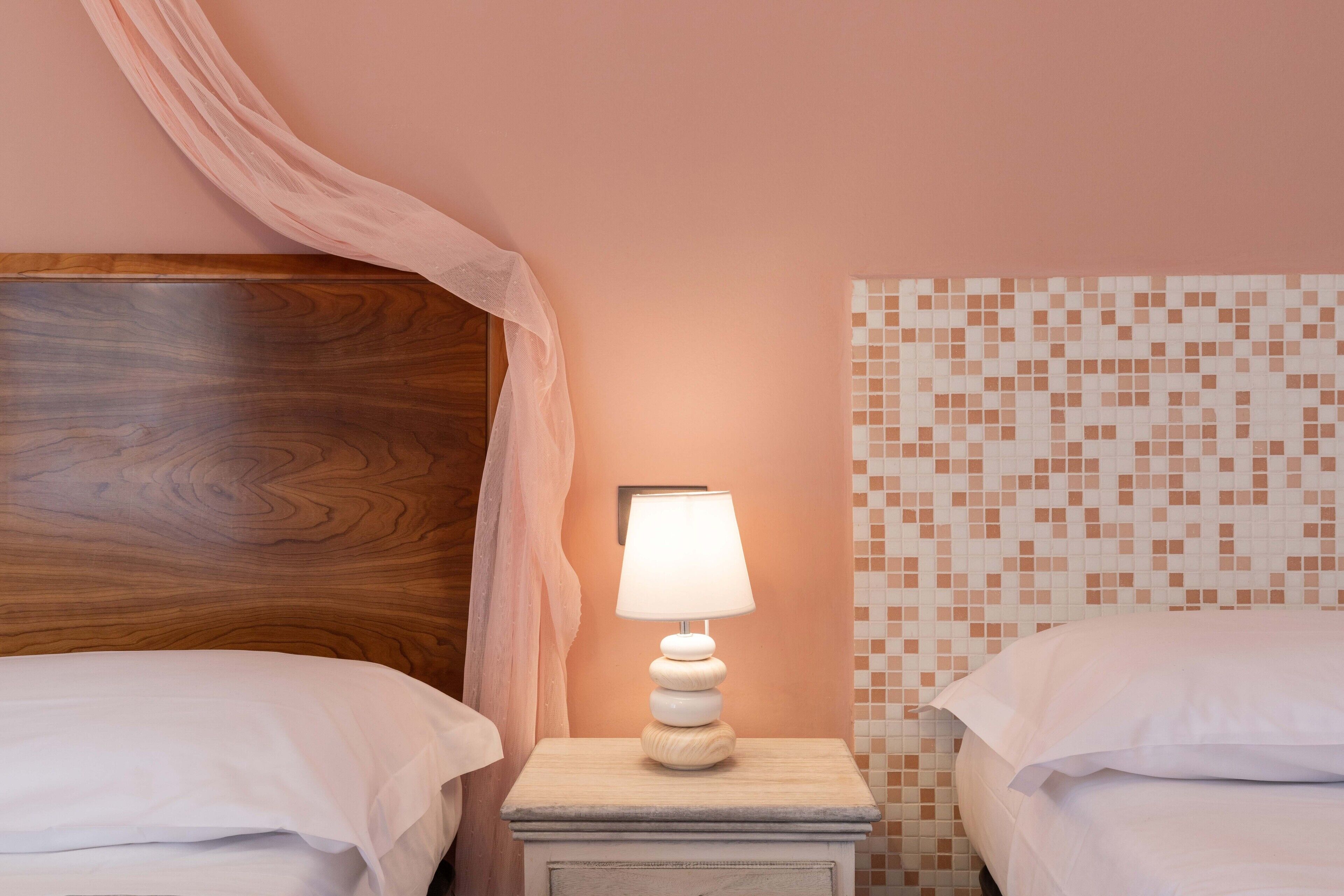 Un dormitorio con dos camas, una mesita de noche con lámpara, un cabecero de madera y una pared rosa con diseño de mosaico.
