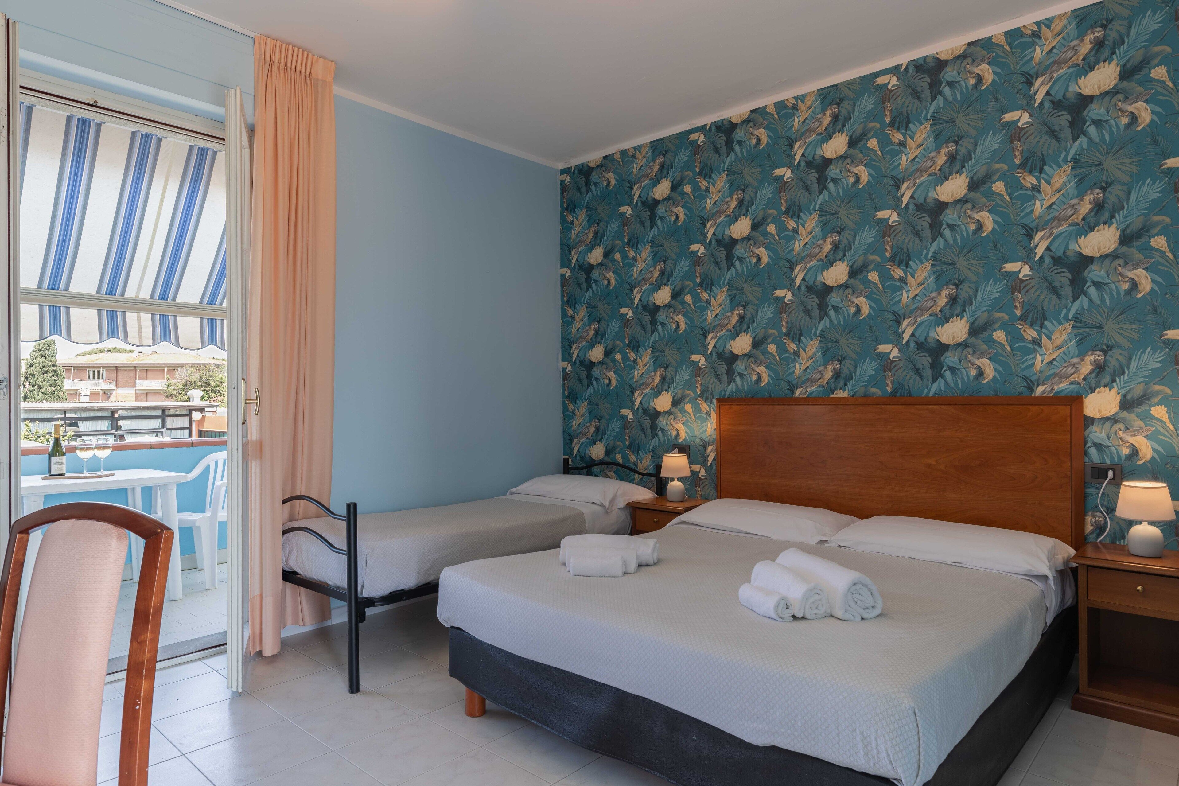 Una camera d'albergo con due letti singoli, una testiera in legno, un tavolino con una sedia e vista all'esterno tramite un'ampia finestra con tende.