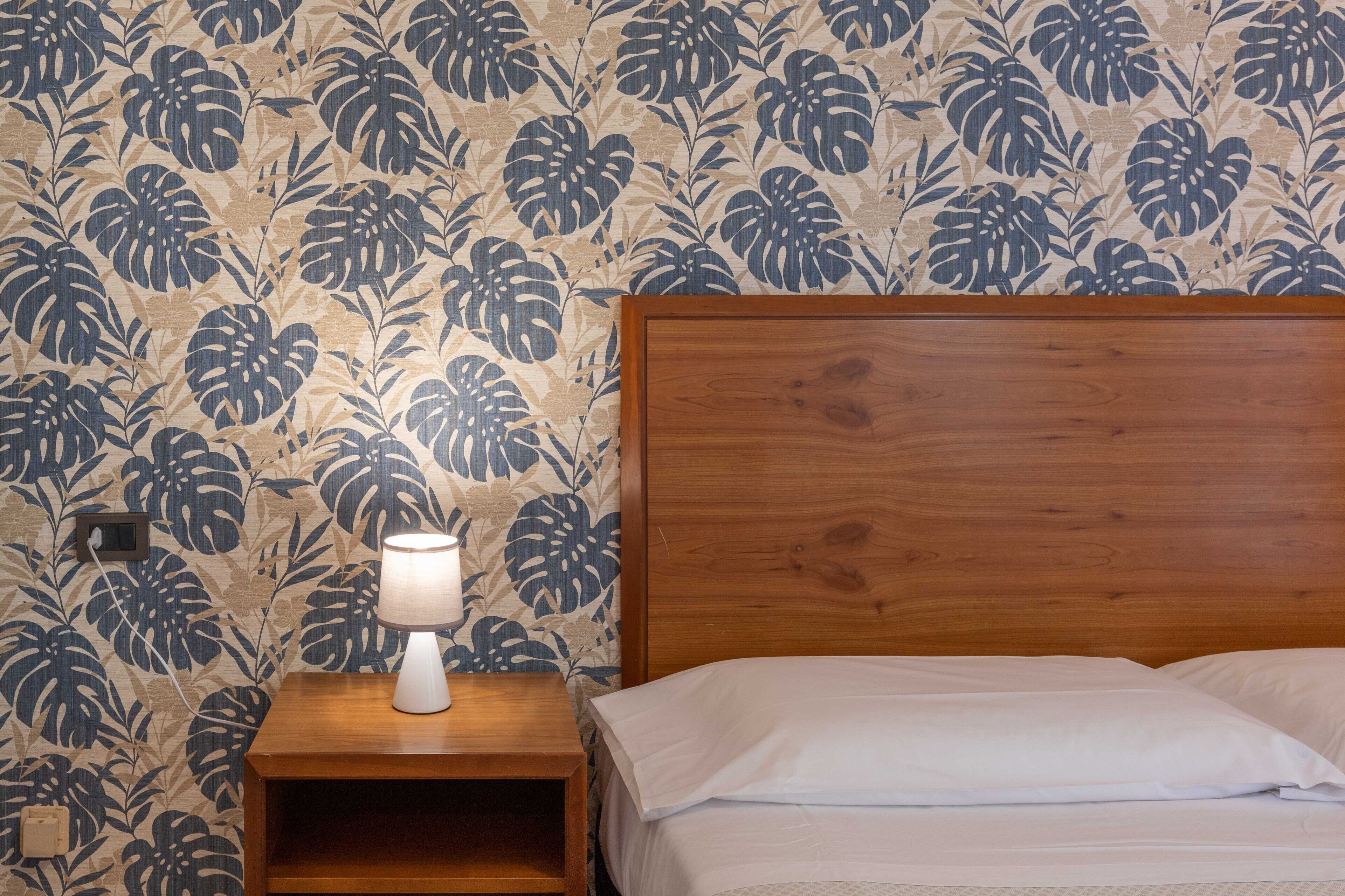 A bedroom with a wooden headboard, a bedside table with a lamp, and a floral wallpaper pattern.