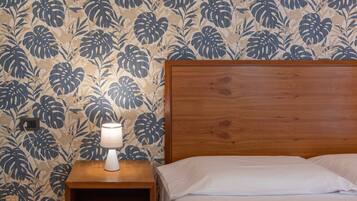 A bedroom with a wooden headboard, a bedside table with a lamp, and a floral wallpaper pattern.