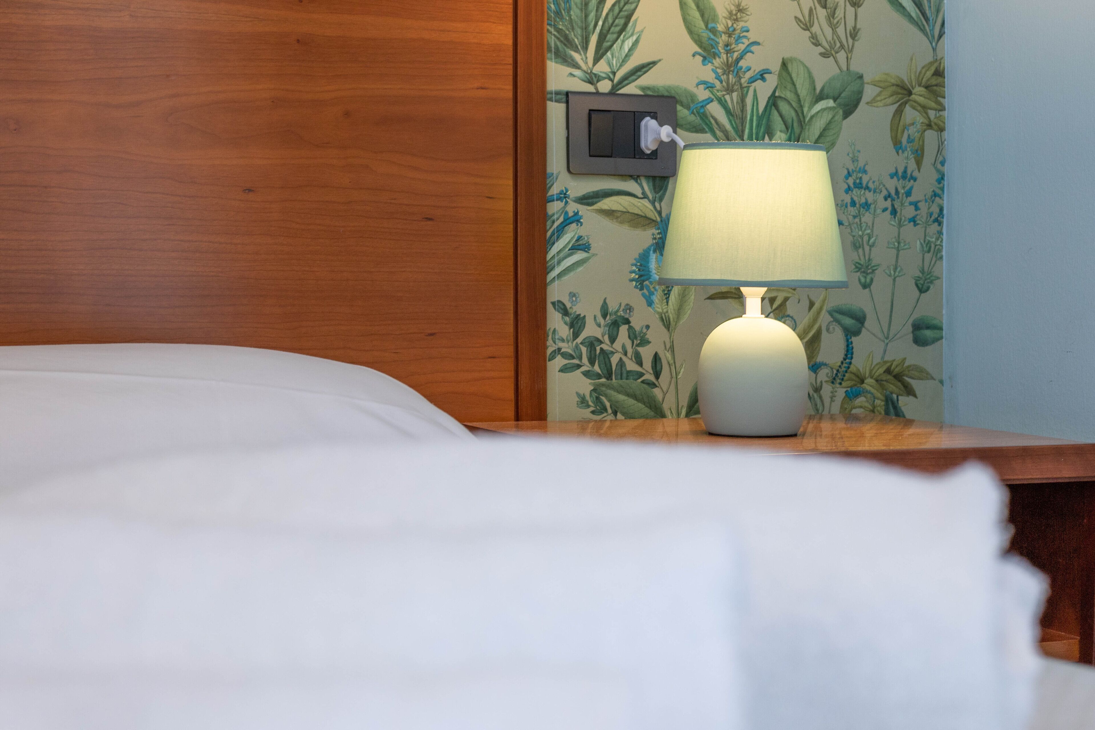 A neatly made bed with white linens, a white bedside lamp, and a night switch on the wall.
