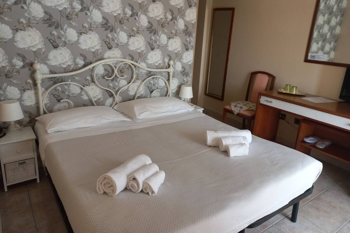 A neatly made bed with white linens and rolled towels, a wooden nightstand with a lamp, a desk with a mirror, and a floral wallpaper pattern.