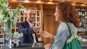 A reception area with a counter, a vase with white flowers, and a bar in the background. - Hotel Riva Etrusca (Bibbona)