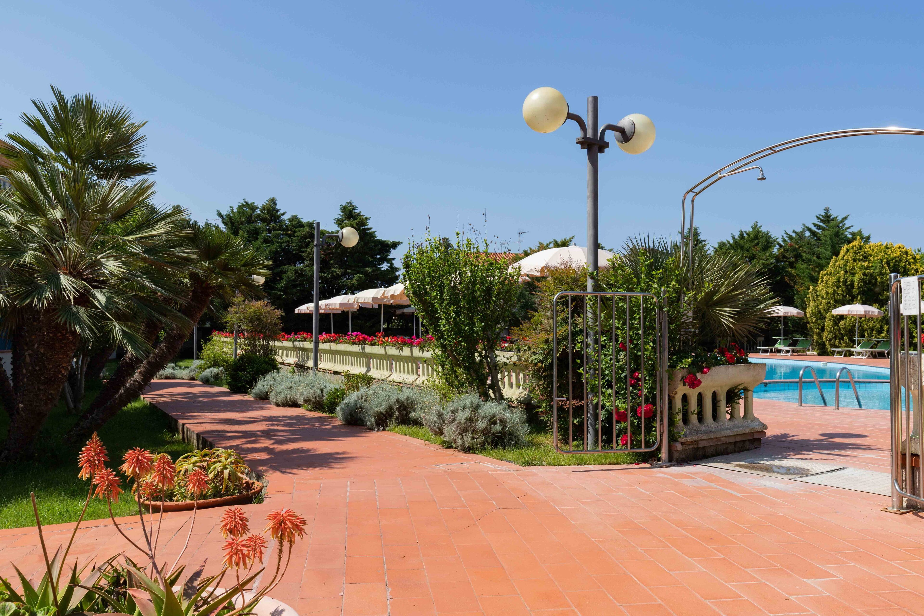 Ein gepflasterter Weg führt zu einem Poolbereich mit einer Fontäne, umgeben von üppiger Vegetation und Palmen.