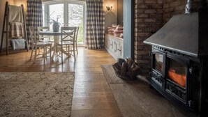 A cozy living room with a fireplace, wooden flooring, a dining area with a table and chairs, and a view through a window with curtains. - Highfield - 3 Bedroom Holiday Home - Llanmadoc (Swansea)