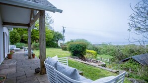 A covered patio with a white bench, a stone pathway, and a view of a lush garden and hills in the distance. - Highfield - 3 Bedroom Holiday Home - Llanmadoc (Swansea)