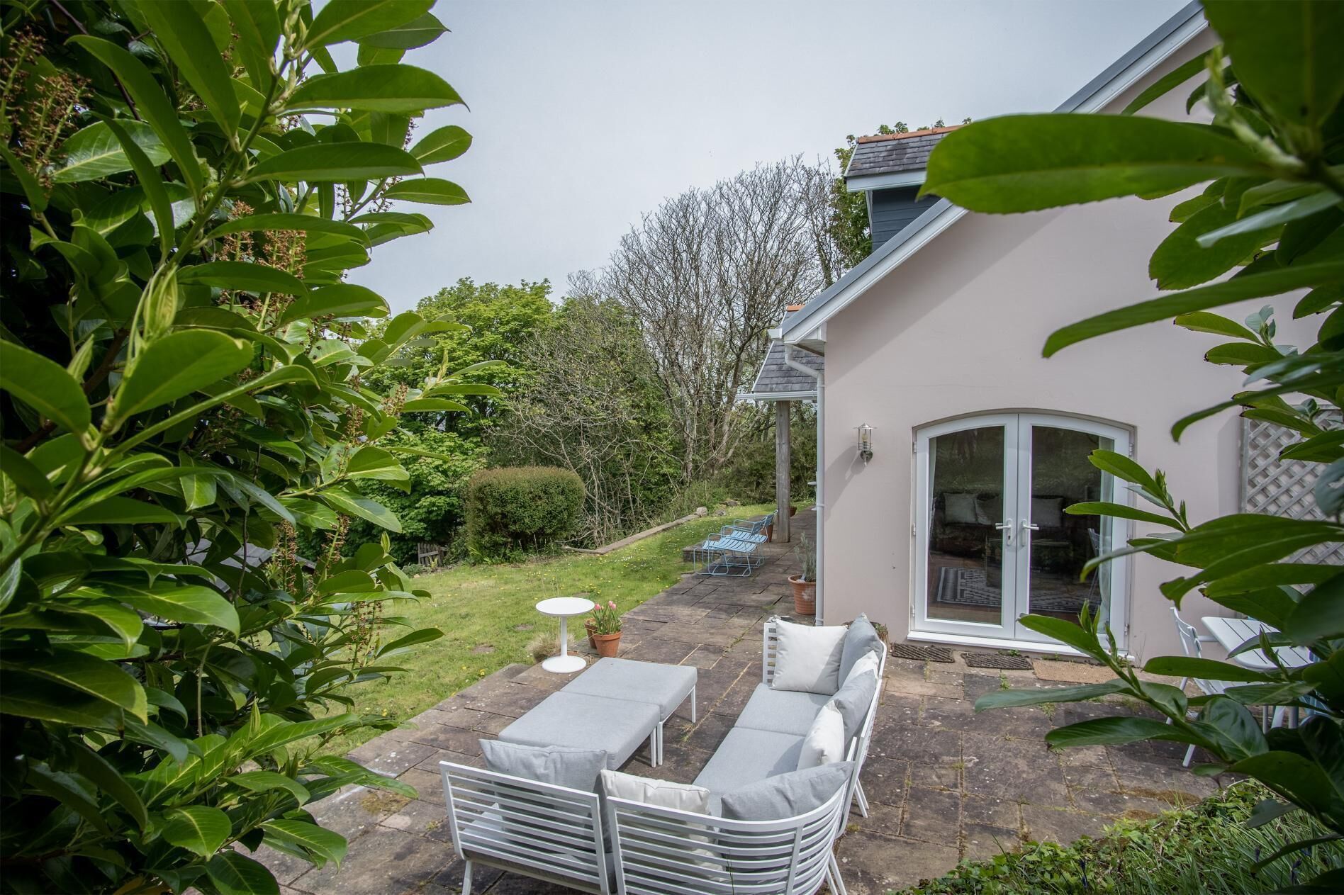 Eine Terrasse mit Tisch und zwei Liegestühlen, ein kleiner Garten und ein Haus mit Glastür.