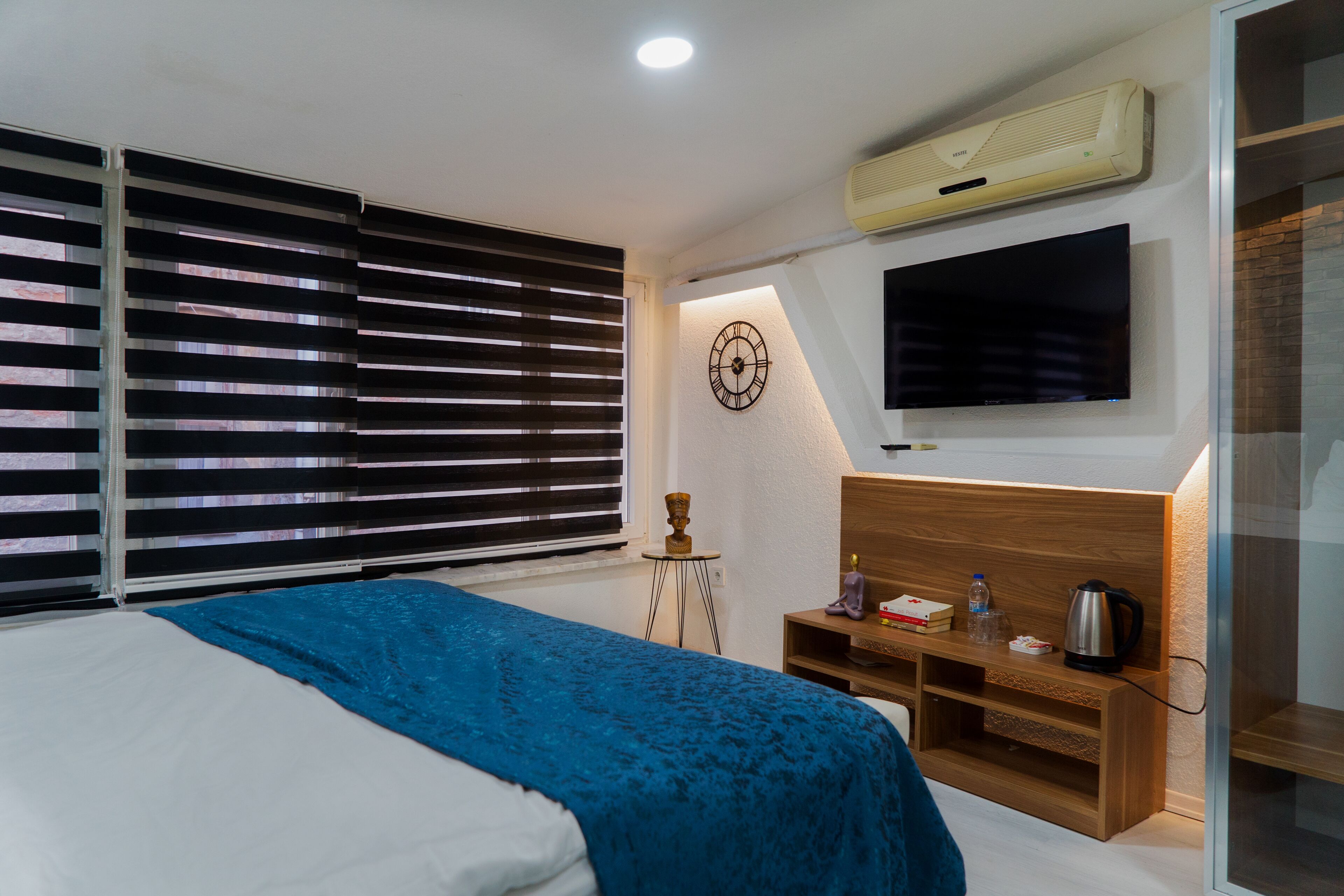 A modern bedroom with a bed, a TV, a clock, and a small table with a kettle and books.