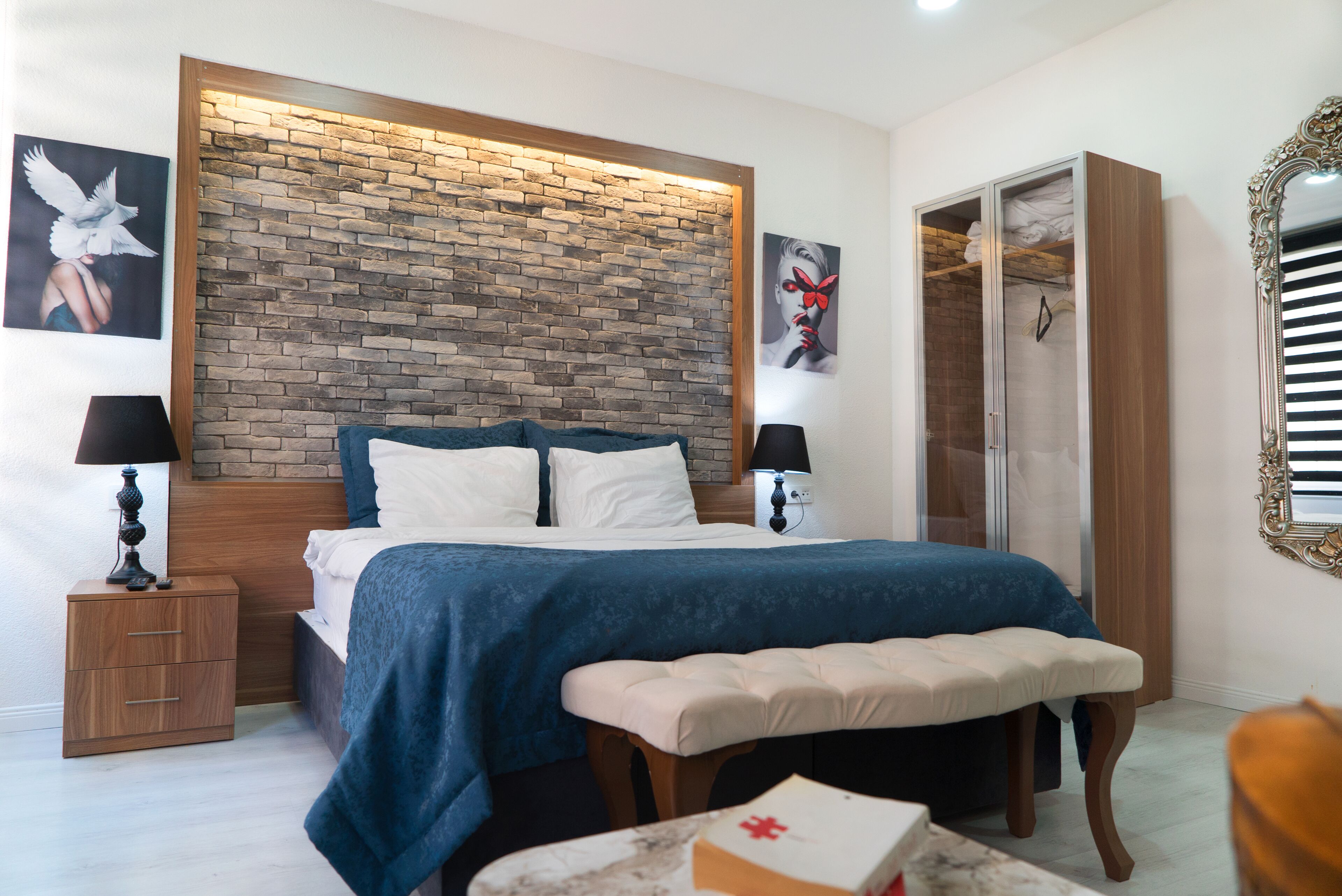 A bedroom with a brick-patterned headboard, a bed with blue bedding, a bedside table with a lamp, a bench, and a wardrobe.