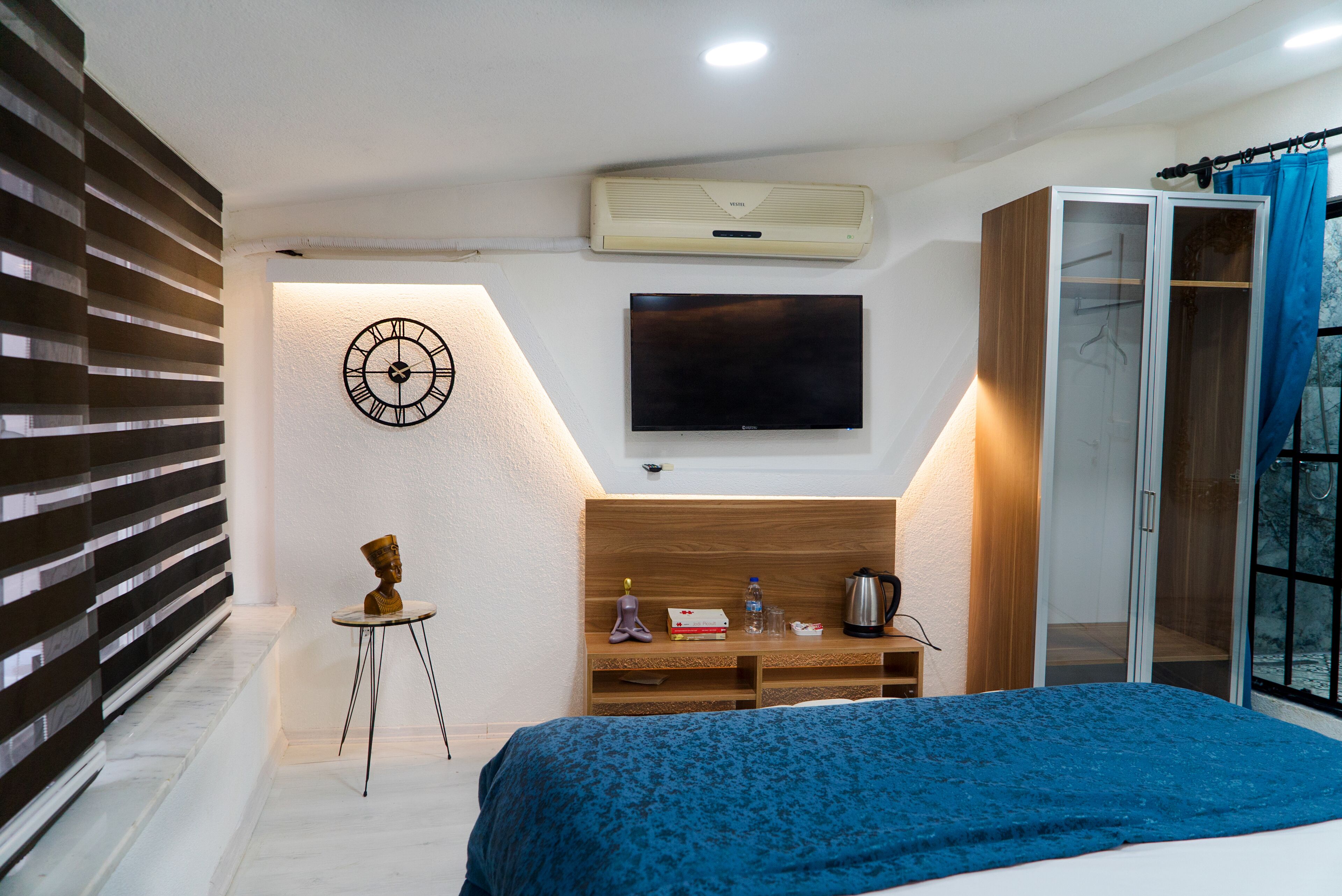 A bedroom with a bed, a TV, a wardrobe, a small table with items, and a wall clock.