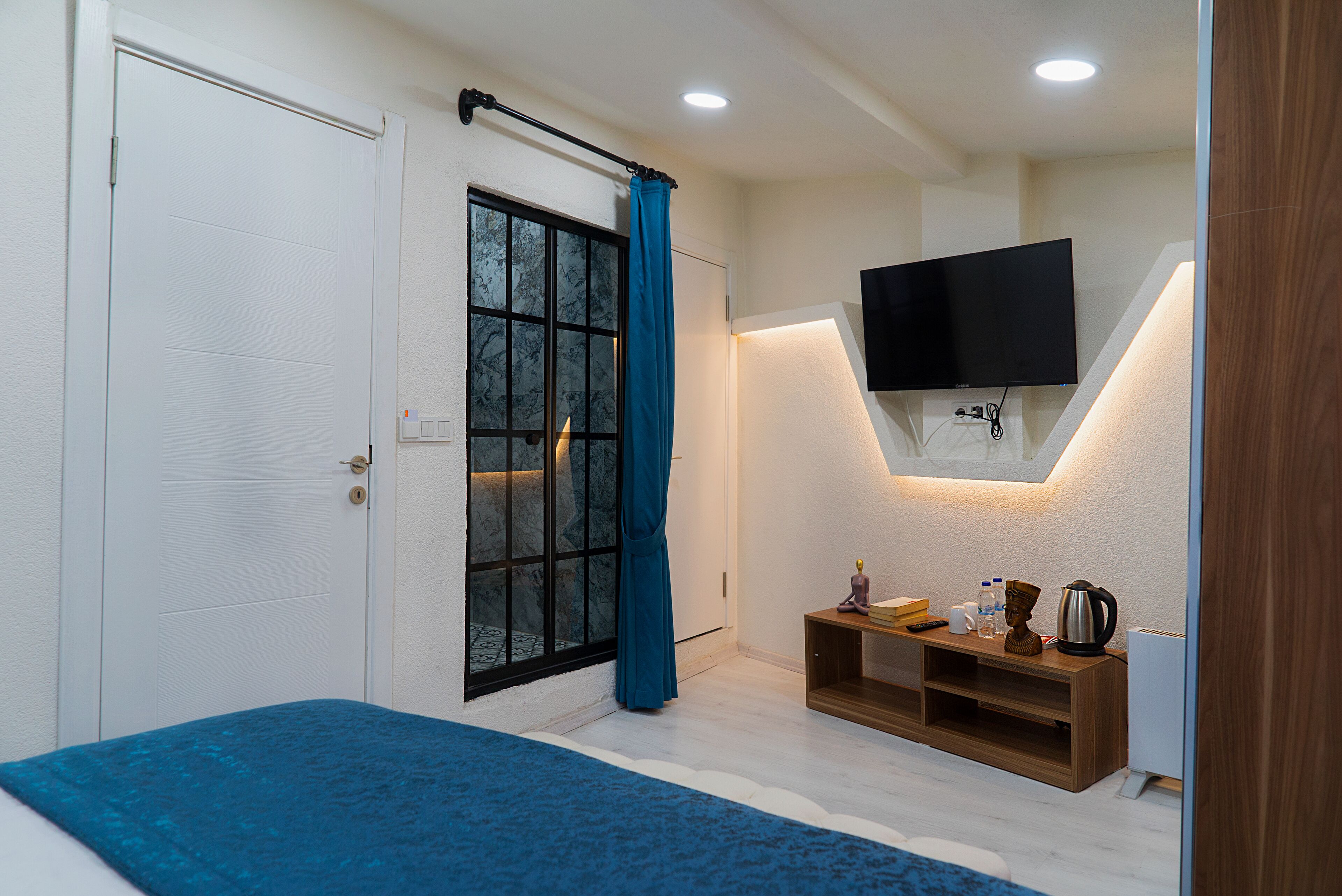 A bedroom with a bed, a TV mounted on the wall, a window with curtains, and a small wooden side table with a kettle and other items.