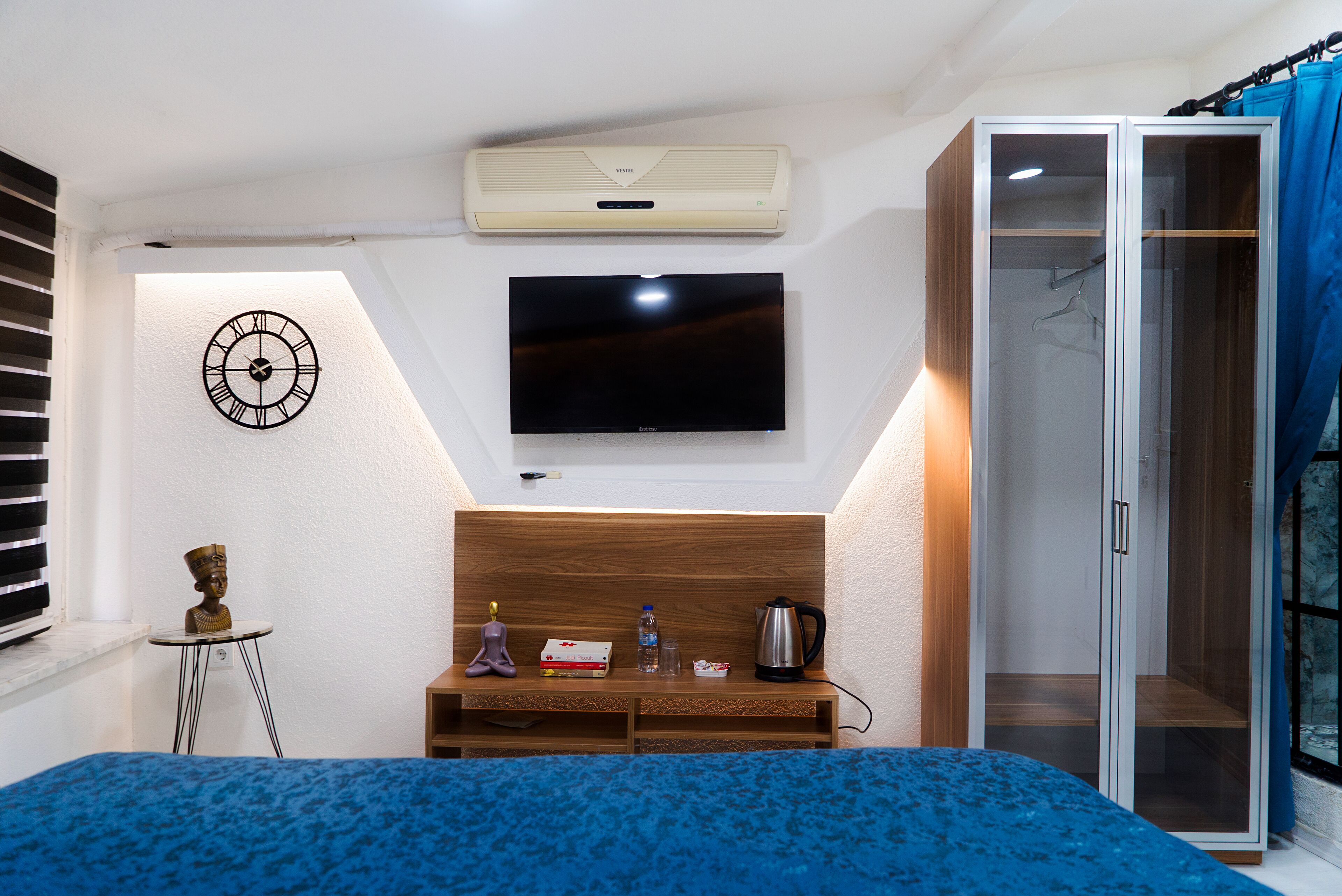 Un dormitorio con cama, televisor, armario, una mesita con objetos y un reloj de pared.