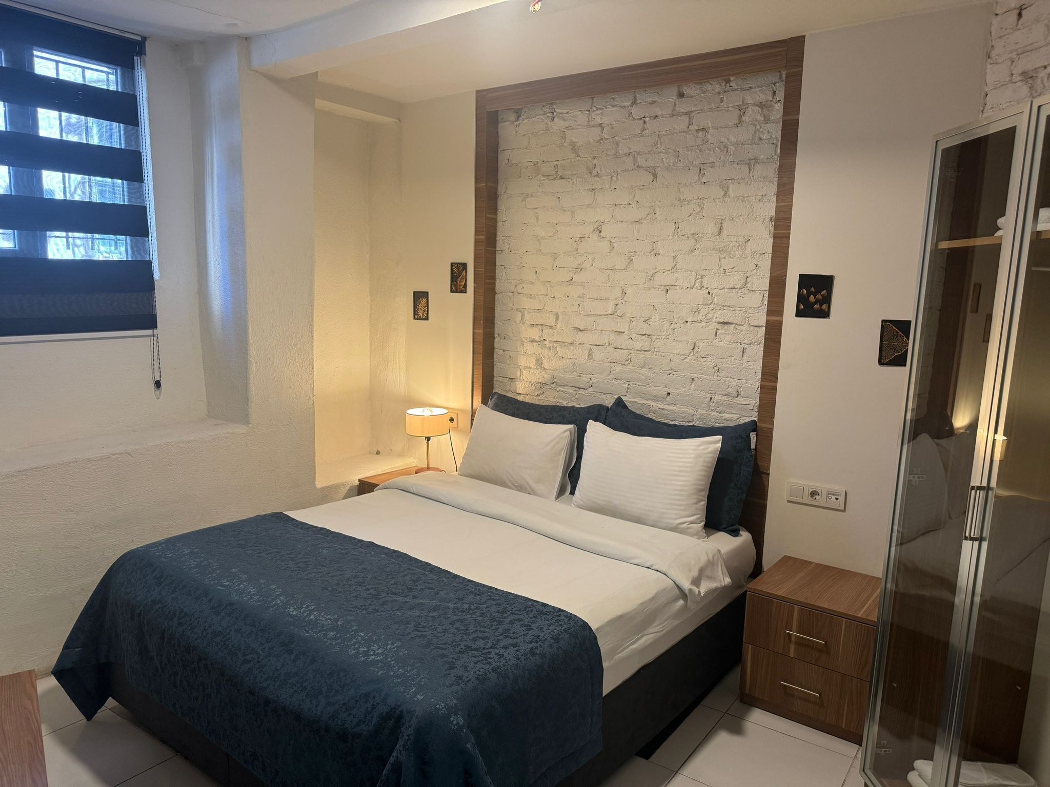 A bedroom with a bed, a nightstand, a wall-mounted lamp, a window with blinds, and a shower area.