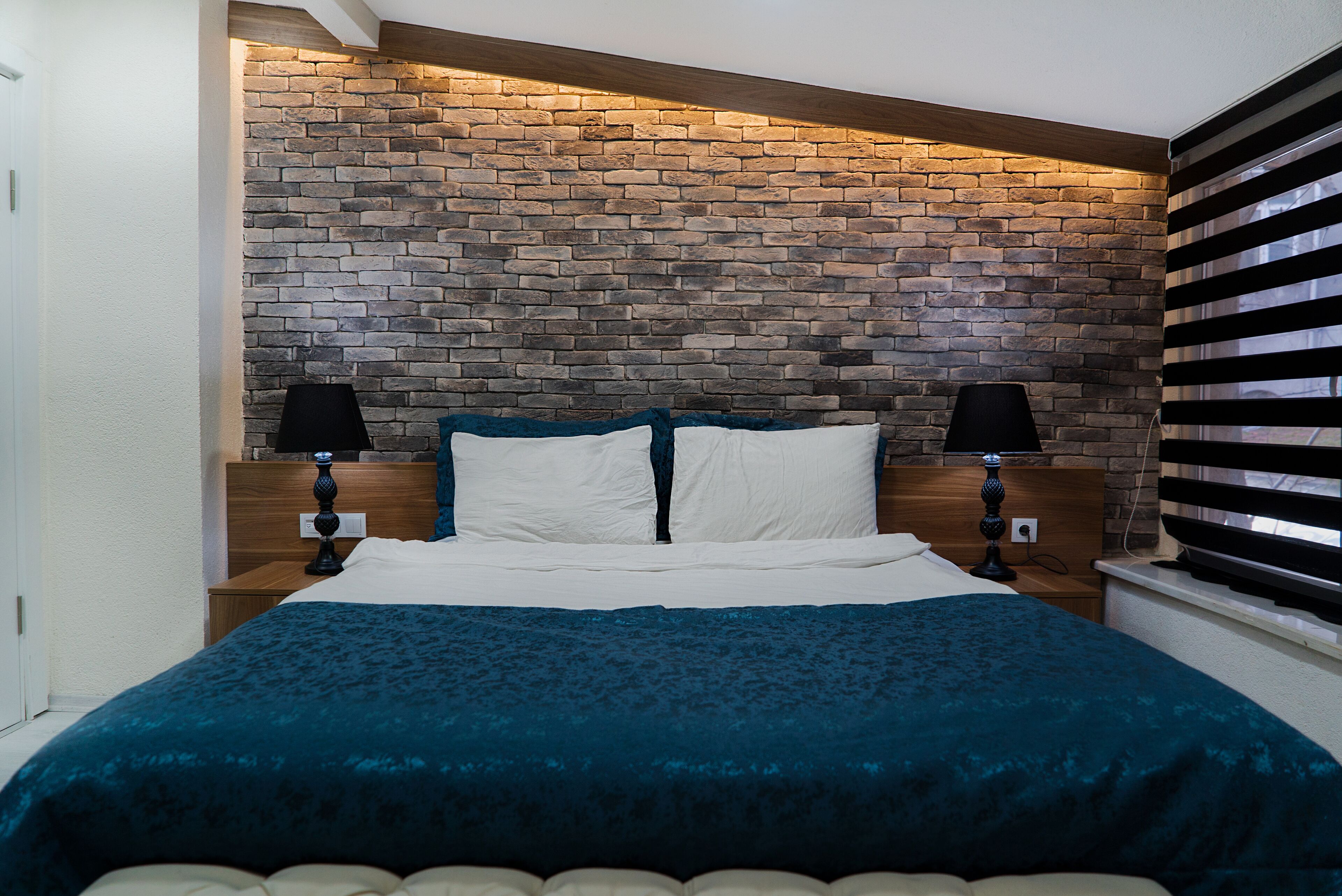 A bedroom with a brick-patterned headboard, a bed with a teal bedspread, two bedside lamps, and a window with blinds.